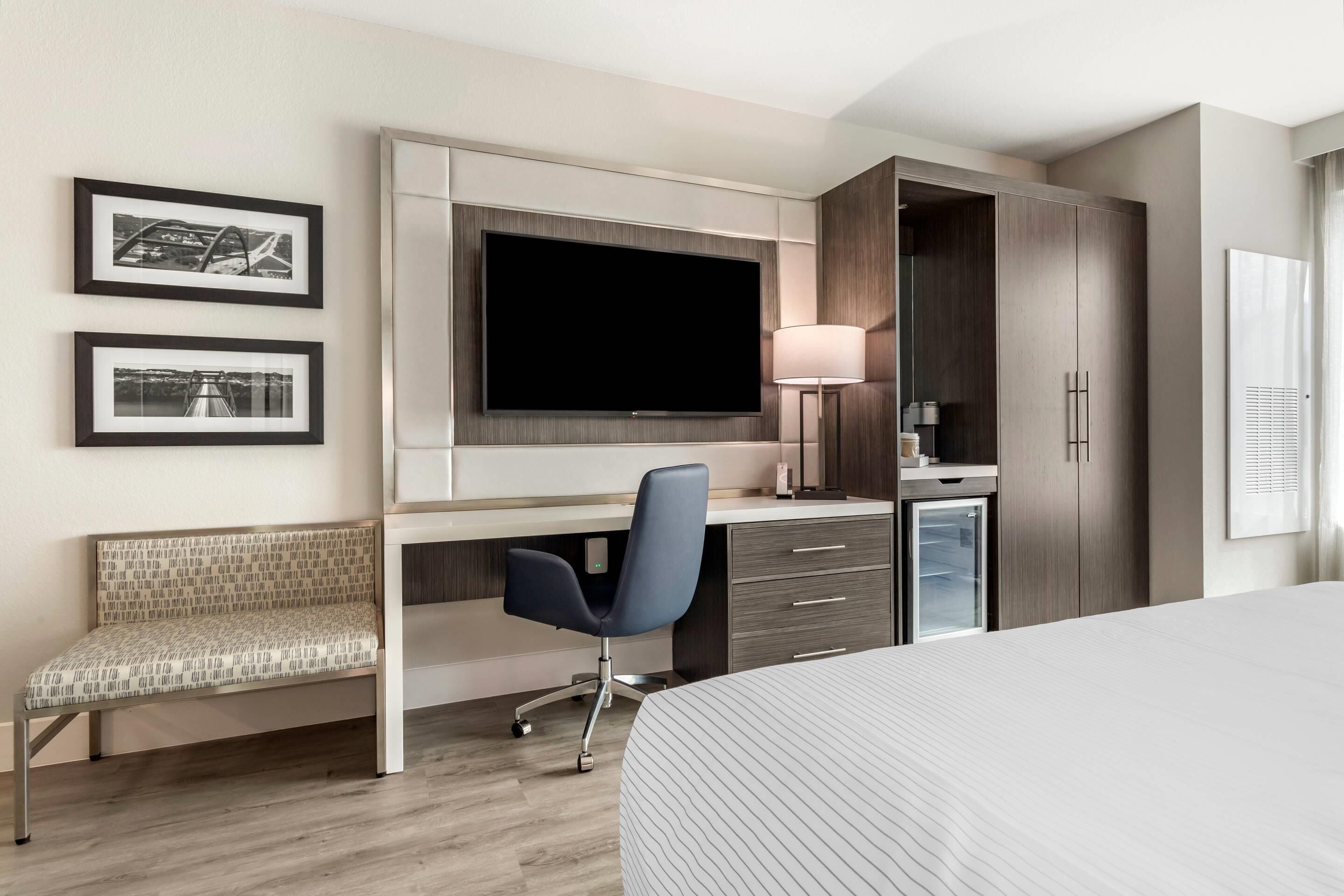 Habitación de hotel con cama, escritorio con silla, televisión, lámpara, armario y dos cuadros en la pared.