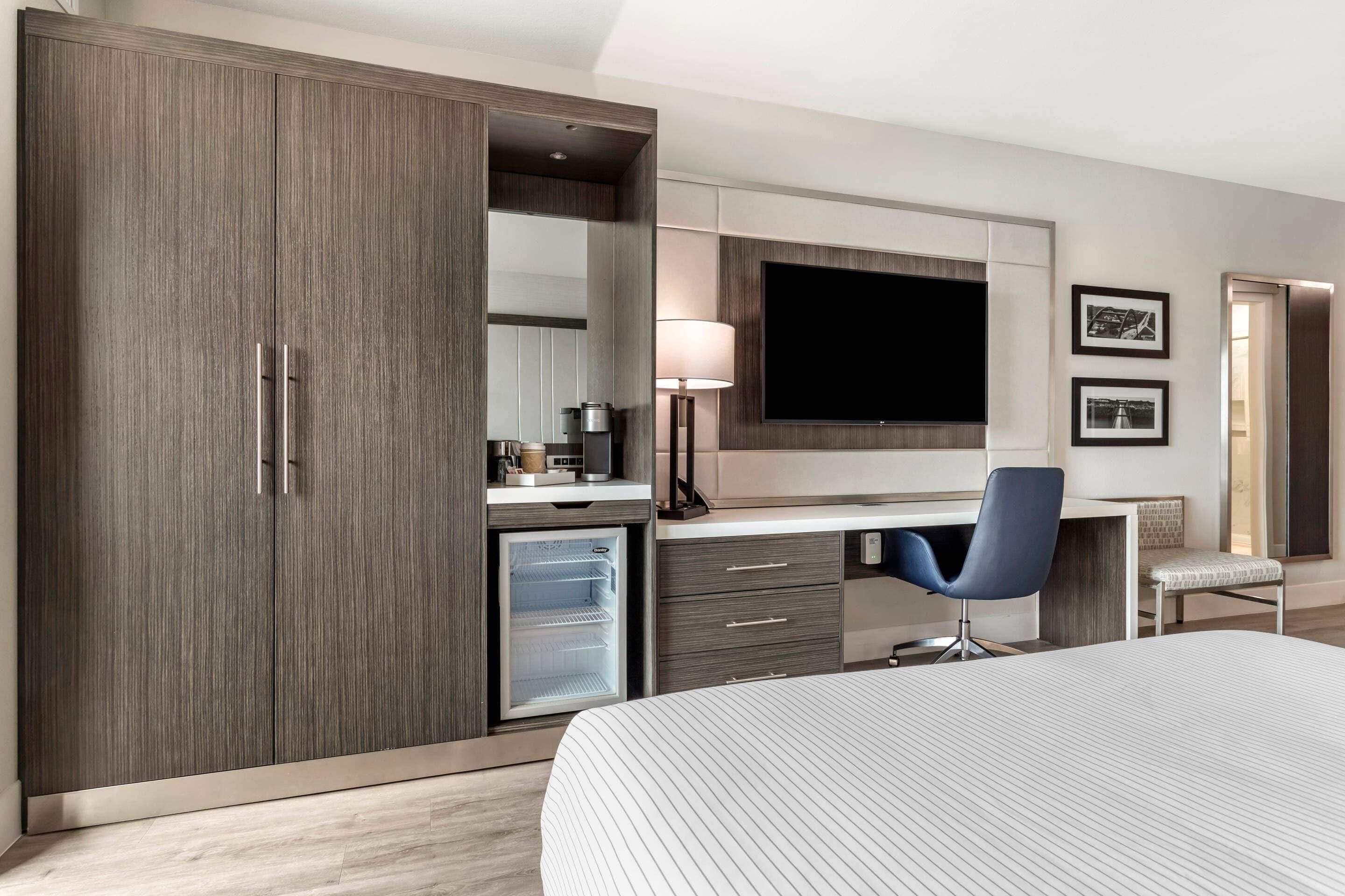 A hotel room with a bed, a desk with a chair, a wardrobe, a television, a mini-fridge, and a lamp.