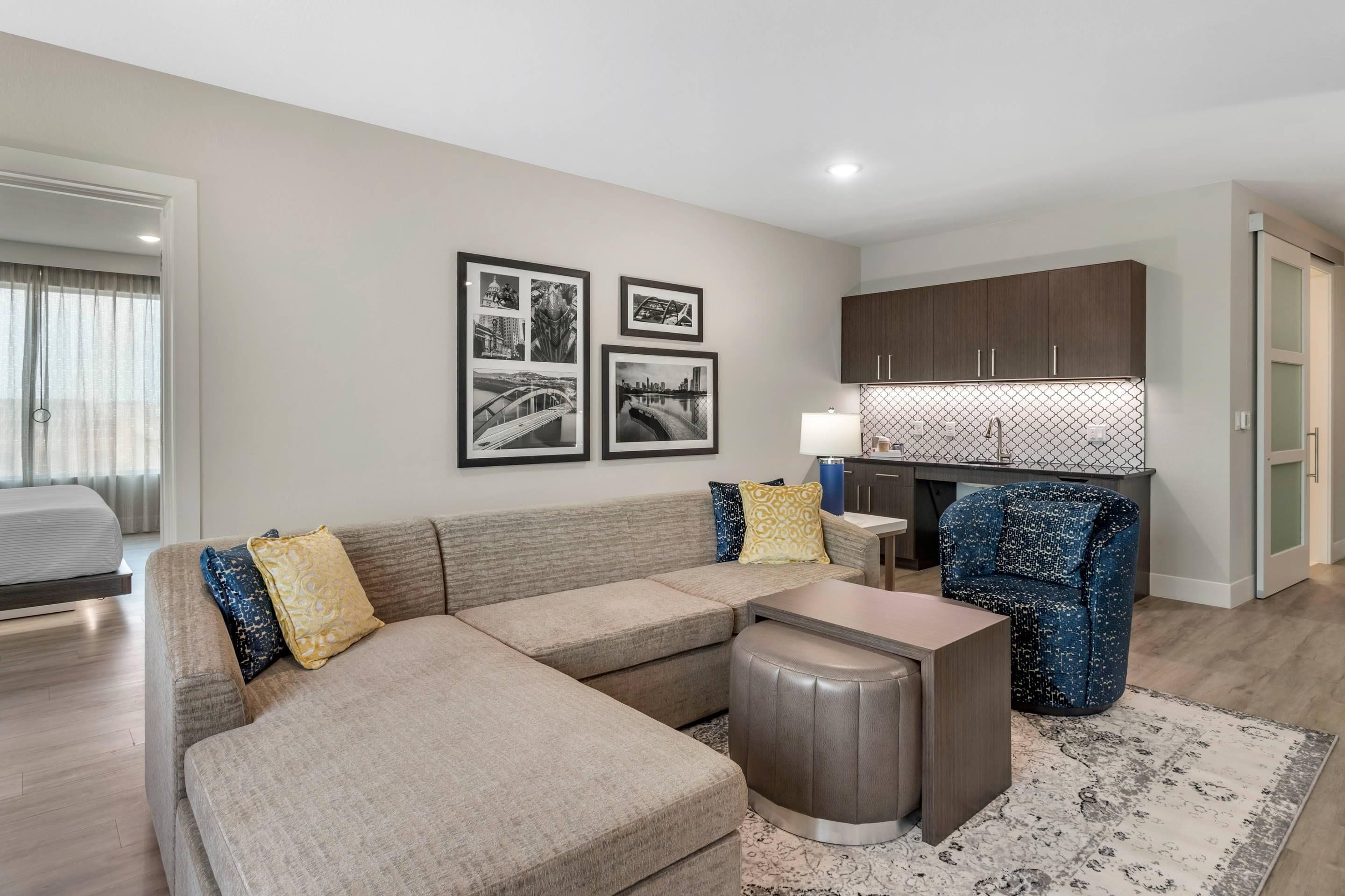 A modern living room with a sectional sofa, a small table, and a patterned rug. There are framed pictures on the wall and a kitchen area with wooden cabinets.