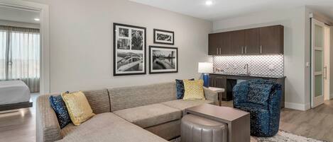 A modern living room with a sectional sofa, a small table, and a patterned rug. There are framed pictures on the wall and a kitchen area with wooden cabinets.