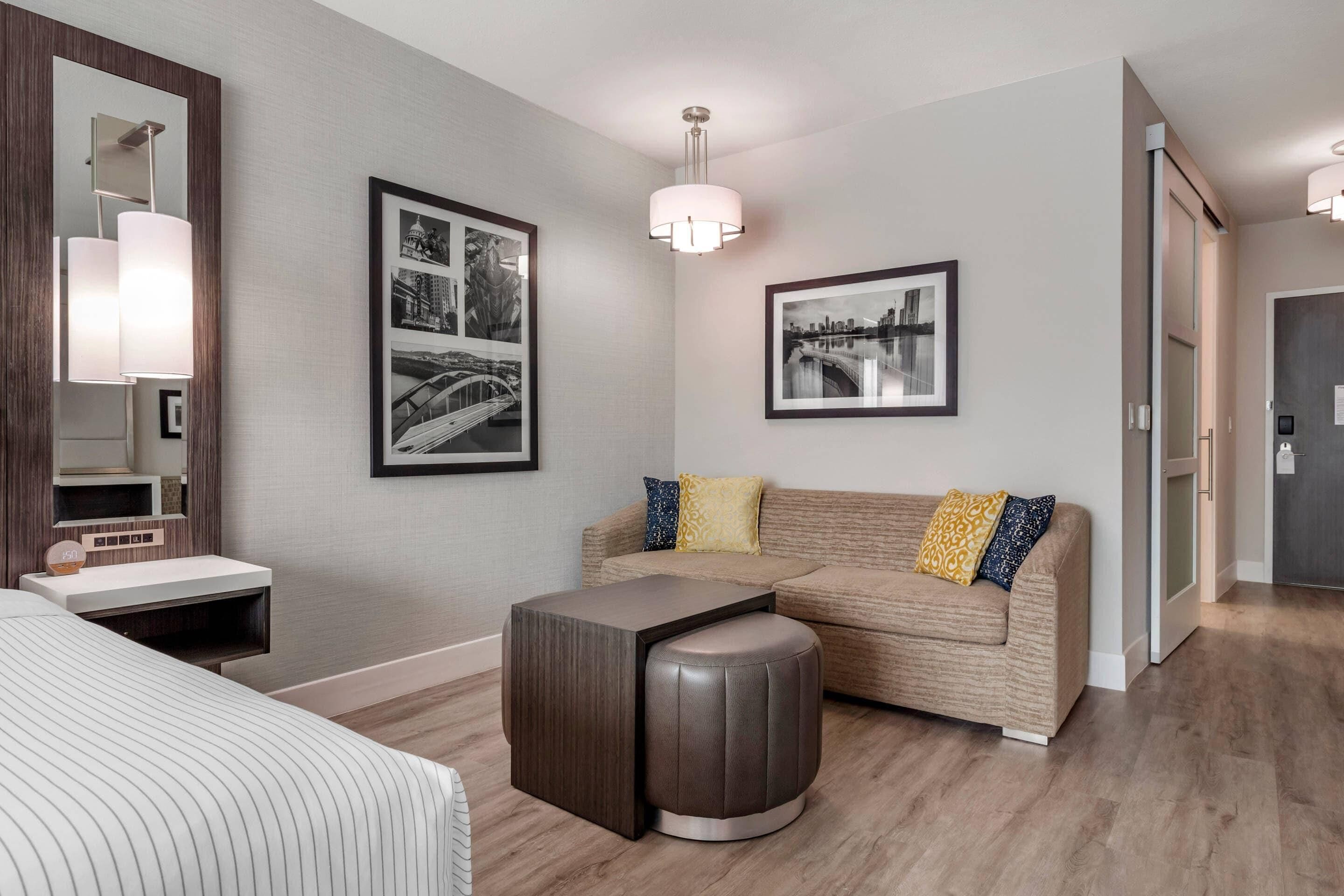 Een moderne hotelkamer met een bed, een bank, een klein tafeltje en een spiegel. Twee ingelijste foto's hangen aan de muur en er is een plafondlamp.