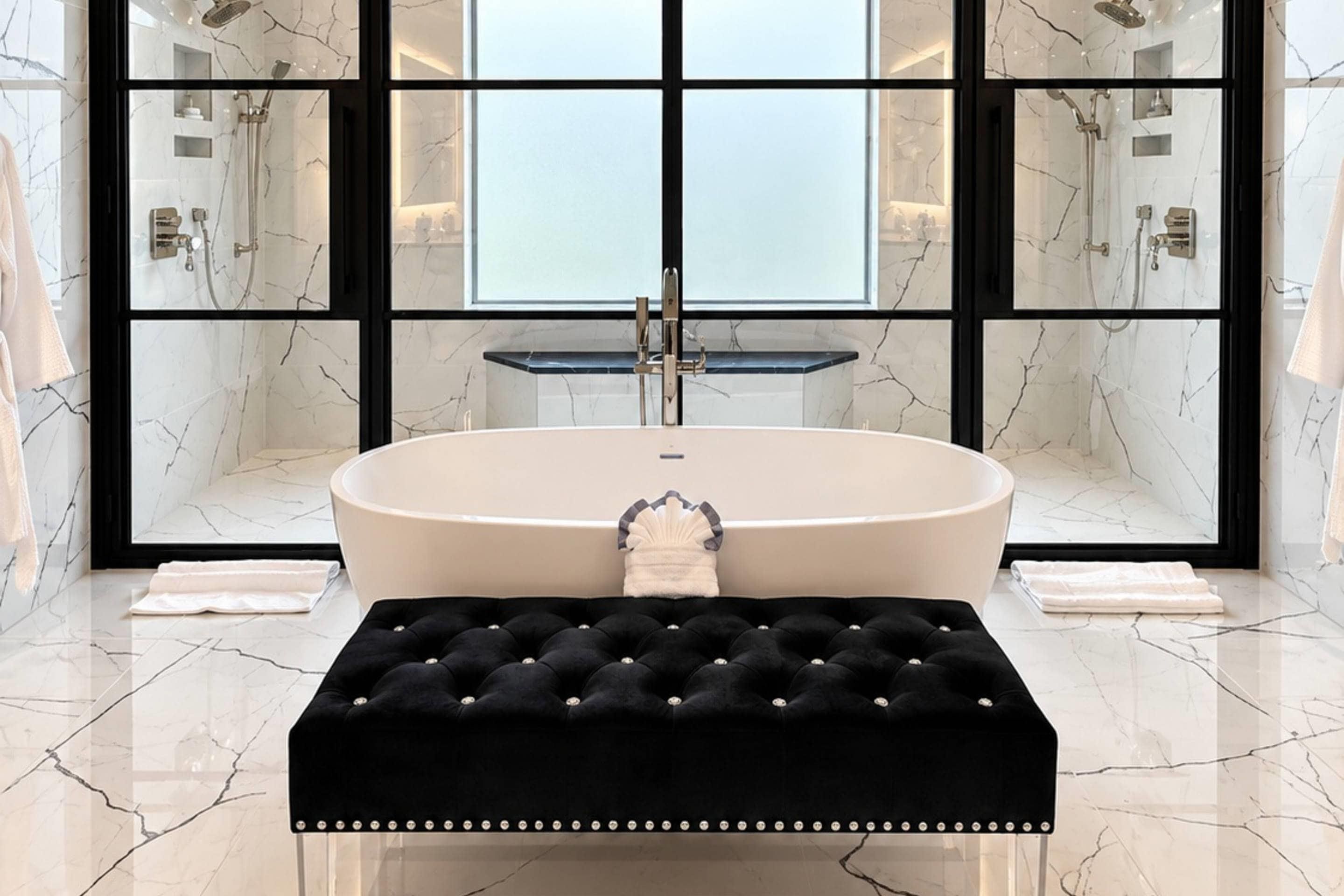 A modern bathroom with a freestanding bathtub, marble walls, and a black tufted bench.