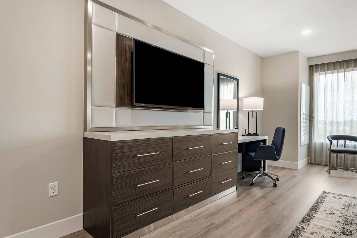 A modern hotel room with a large flat-screen TV mounted on a cabinet, a desk with a chair, and a large window with curtains.