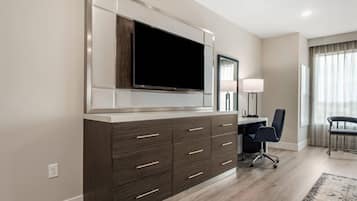 A modern hotel room with a large flat-screen TV mounted on a cabinet, a desk with a chair, and a large window with curtains.