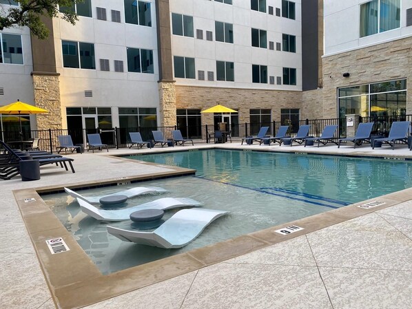 A swimming pool with lounge chairs and umbrellas, surrounded by a building with multiple windows and a stone facade.