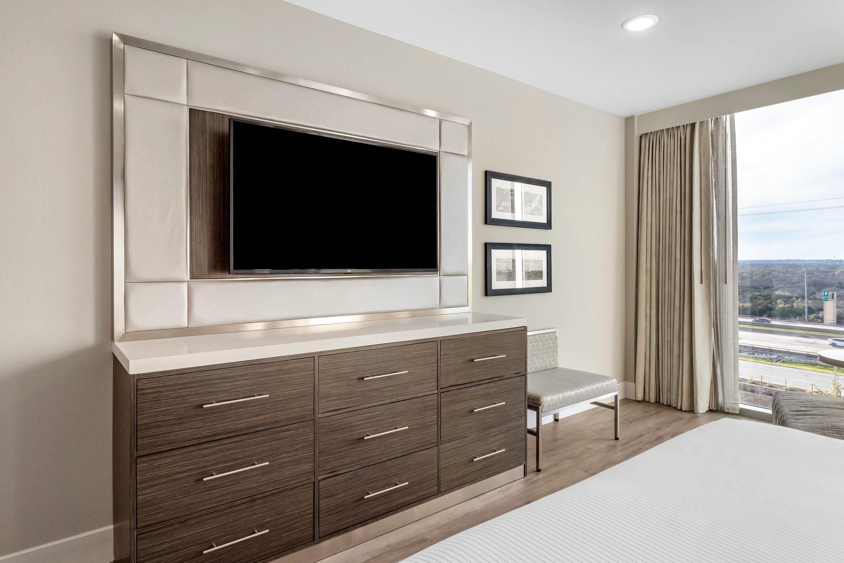 A modern hotel room with a large flat-screen TV, a built-in cabinet with drawers, a bench, and a large window with a city view.