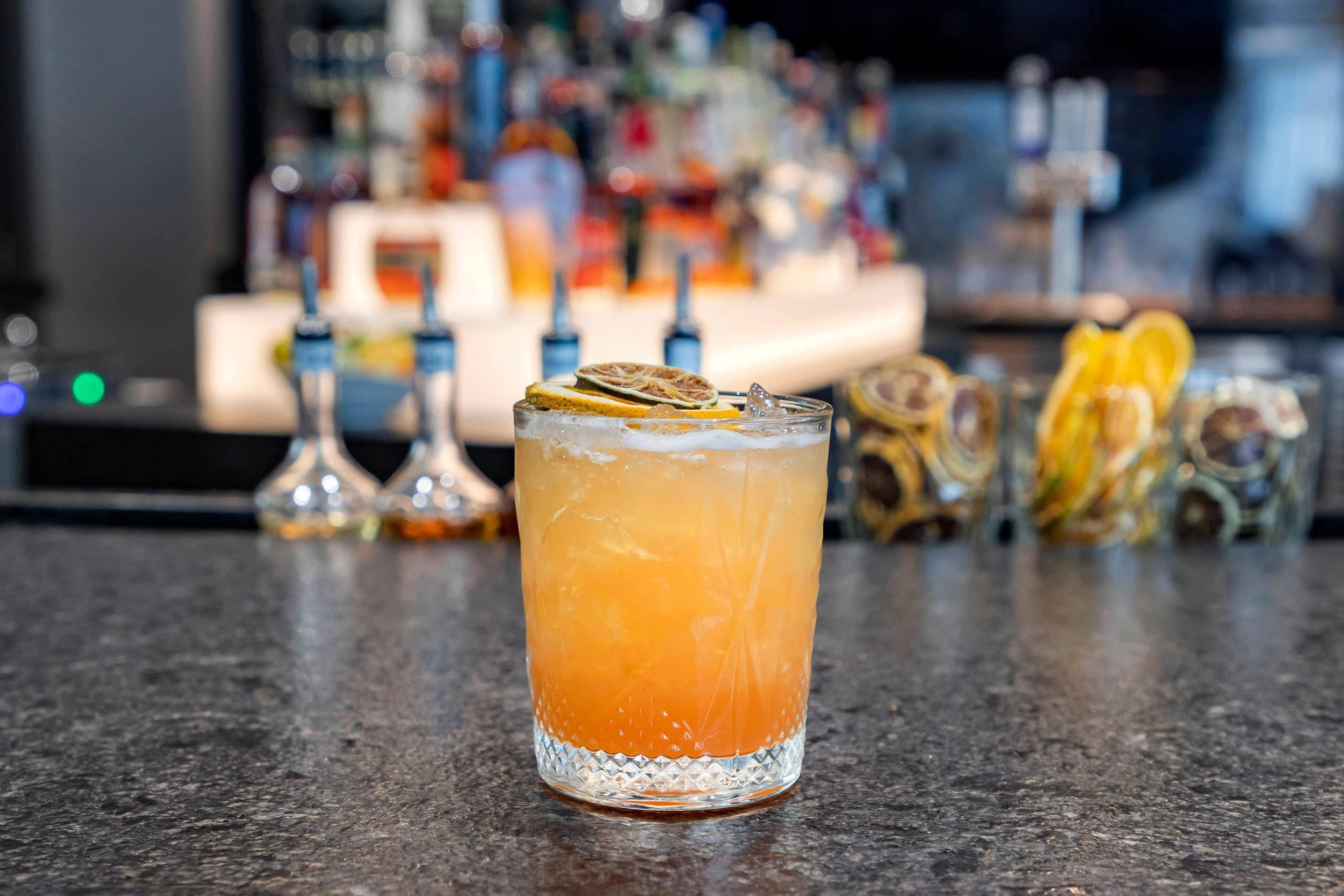 A bar setting with a glass of orange-colored drink garnished with citrus slices and a lemon wheel, with a blurred background of various bottles and bar equipment.