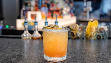 A bar setting with a glass of orange-colored drink garnished with citrus slices and a lemon wheel, with a blurred background of various bottles and bar equipment.