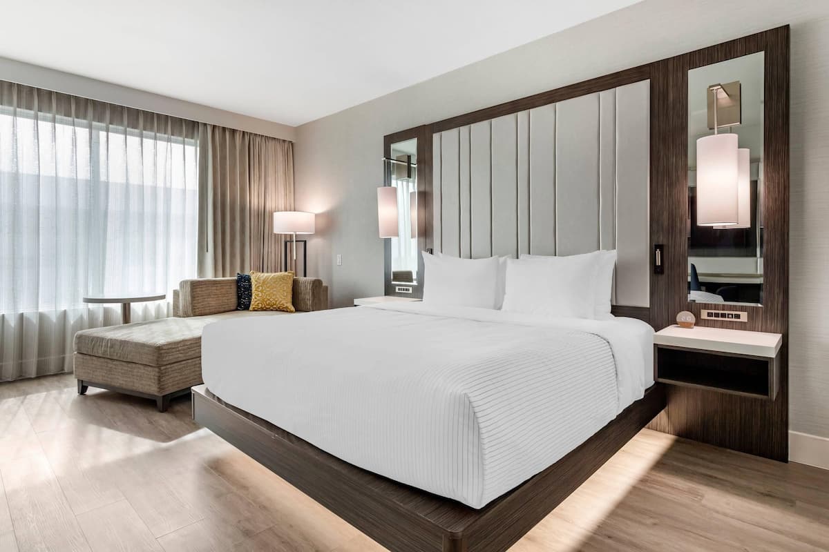 A modern hotel room with a large bed, a bedside table, a chair, a lamp, and a large mirror.