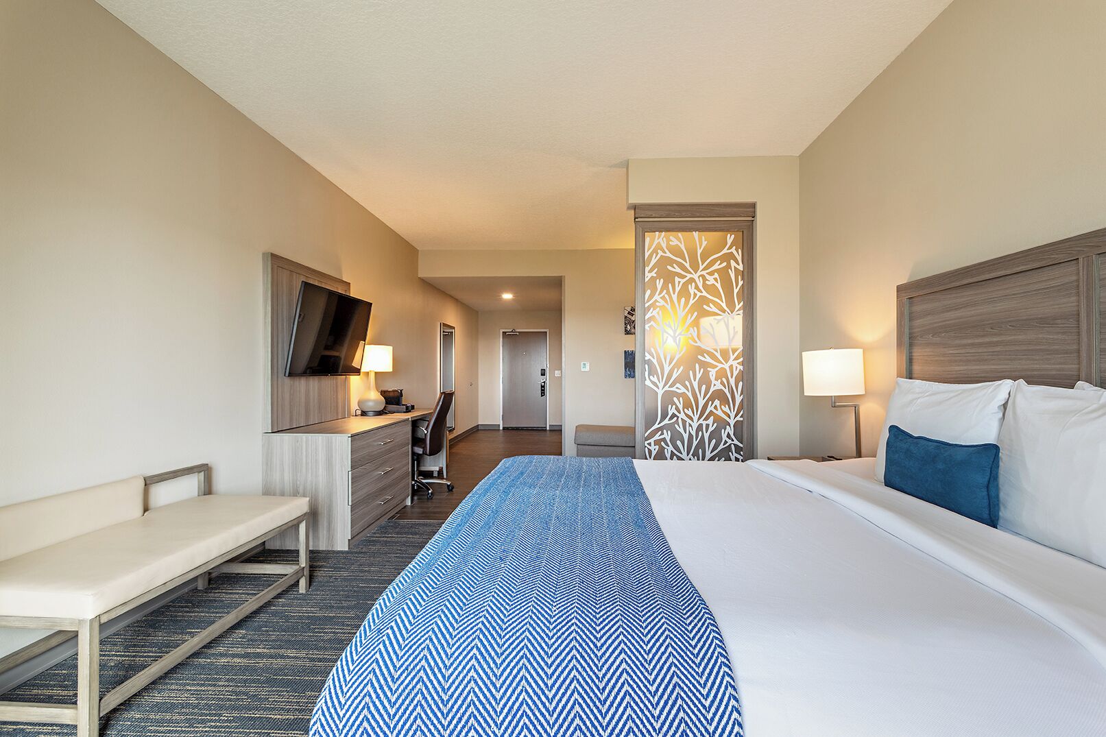A hotel room with a bed, a desk with a chair, a small table, and a TV.