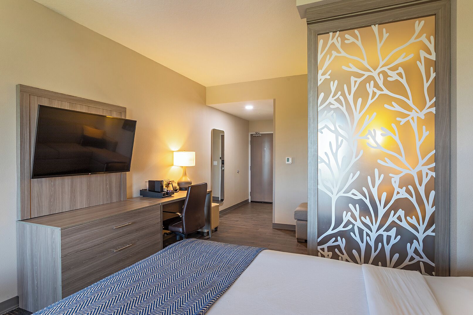 Habitación de hotel con cama, escritorio con silla, televisor, lámpara y un panel decorativo en la pared.
