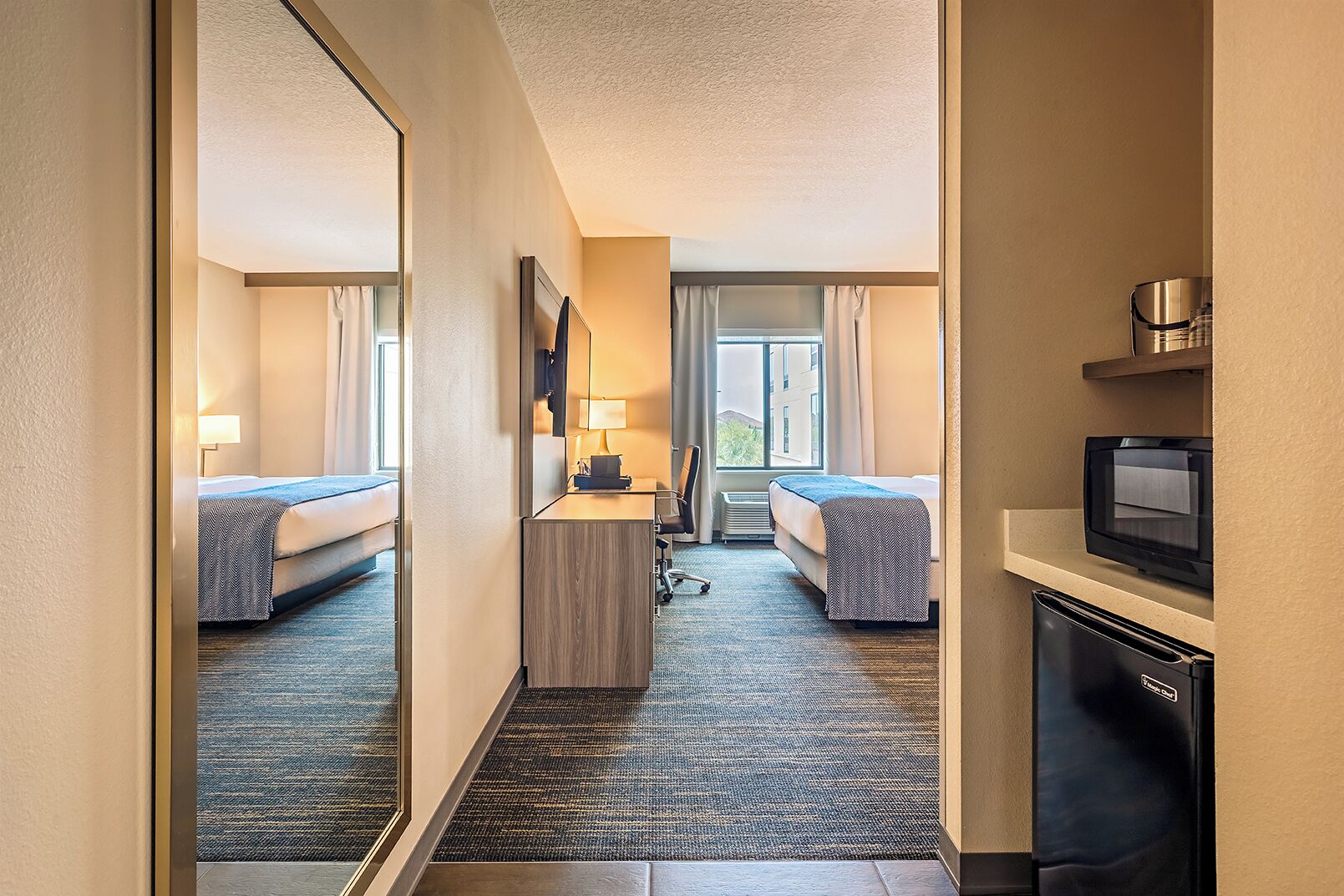 A hotel room with two beds, a desk with a lamp, a television, and a view of the outdoors.