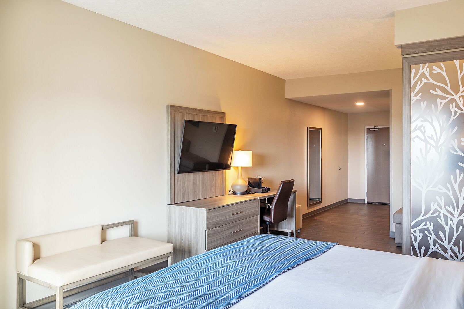 A hotel room with a bed, a desk with a chair, a TV mounted on the wall, and a mirror.