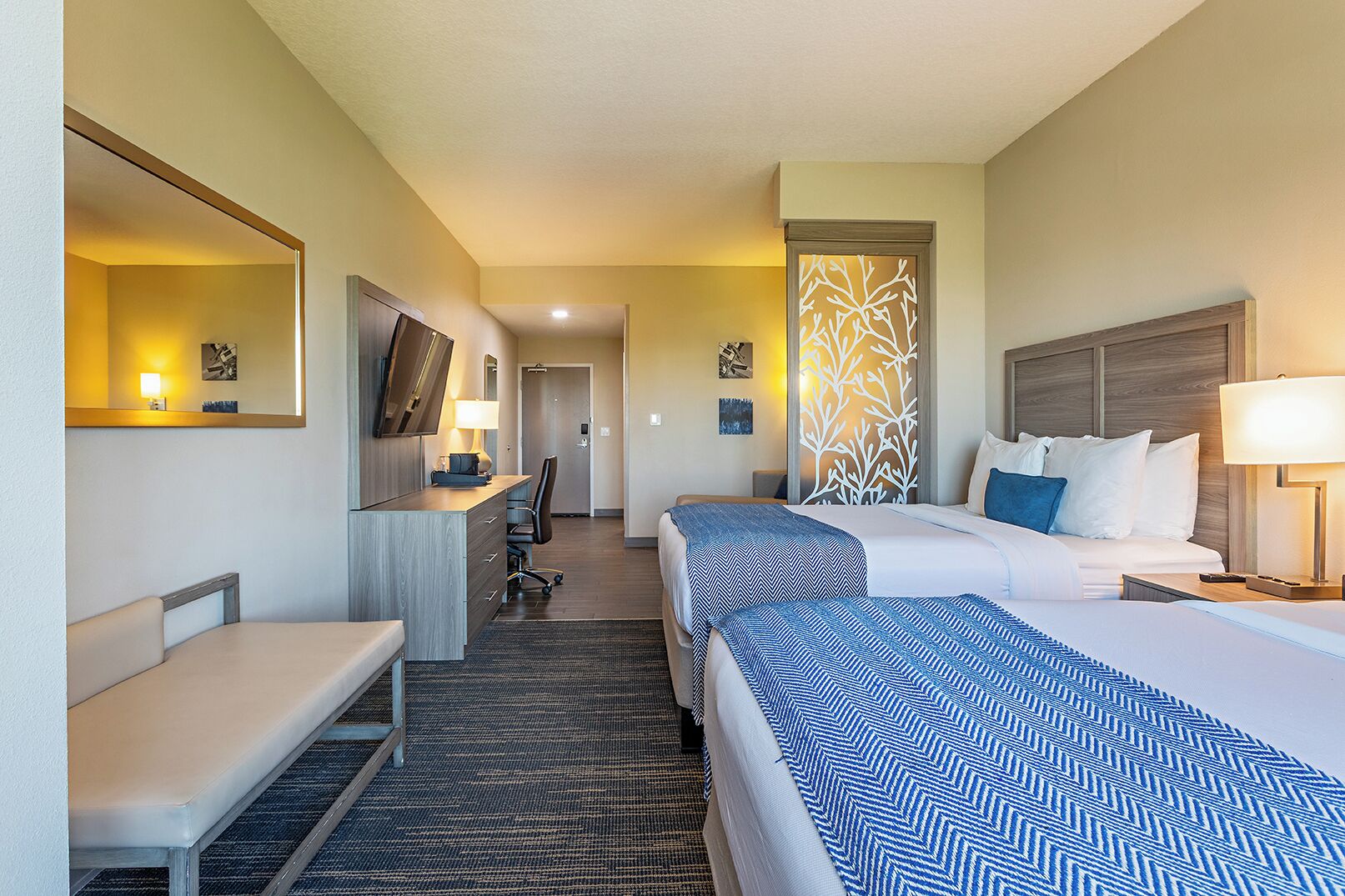 A hotel room with two beds, a desk, a chair, a bench, and a TV.