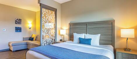 A hotel room with a large bed, a sofa, a nightstand with a lamp, and a decorative wall panel.
