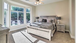 Une chambre avec une grande fenêtre, un ventilateur de plafond, un lit recouvert d’une couvre-lit à motifs, une table de chevet avec une lampe et un sol recouvert d’une moquette.