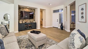 Un salon spacieux avec un mur d’accent en pierre, un téléviseur à écran plat, un parquet en bois foncé et un grand pouf.