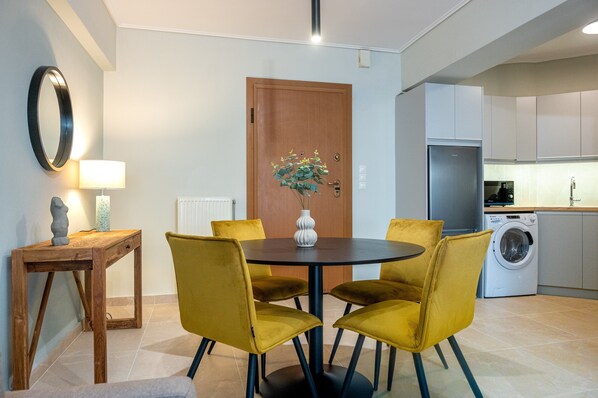 A dining area with a round table, four yellow chairs, a wooden side table with a lamp, a vase with flowers, and a wooden door with a plant decoration.