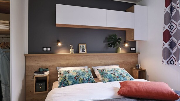 Ein modernes Schlafzimmer mit einem Holzkopfteil, zwei gleichfarbigen floralen Kissen, einem roten Akzentkissen, einem Nachttisch mit Lampe und einer kleinen Pflanze.