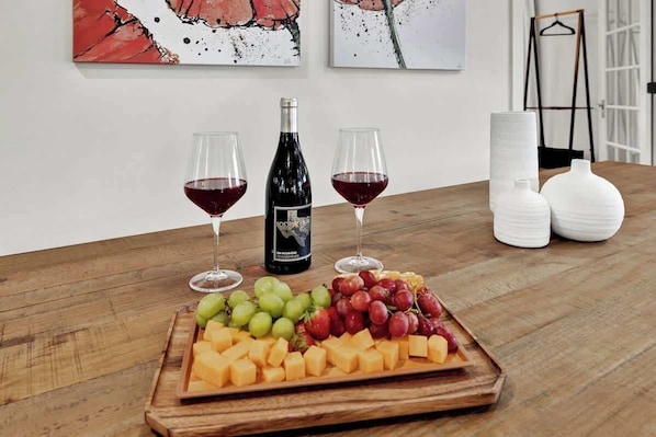 A wooden table with a cheese and fruit platter, two glasses of red wine, a wine bottle, and two white vases.