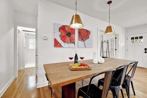 Un comedor con una mesa de madera, sillas negras, dos lámparas colgantes, una bandeja de frutas, copas de vino y un cuadro de estilo pop en la pared.