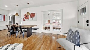 Un salon moderne avec un plancher en bois, un coin repas avec une table et des chaises, un canapé avec des coussins et une grande fenêtre donnant sur une chambre.