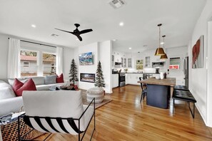 A modern living room with a sofa, a coffee table, and a fireplace. There is a dining area with a table and chairs. The kitchen is visible in the background with white cabinets and a large island.