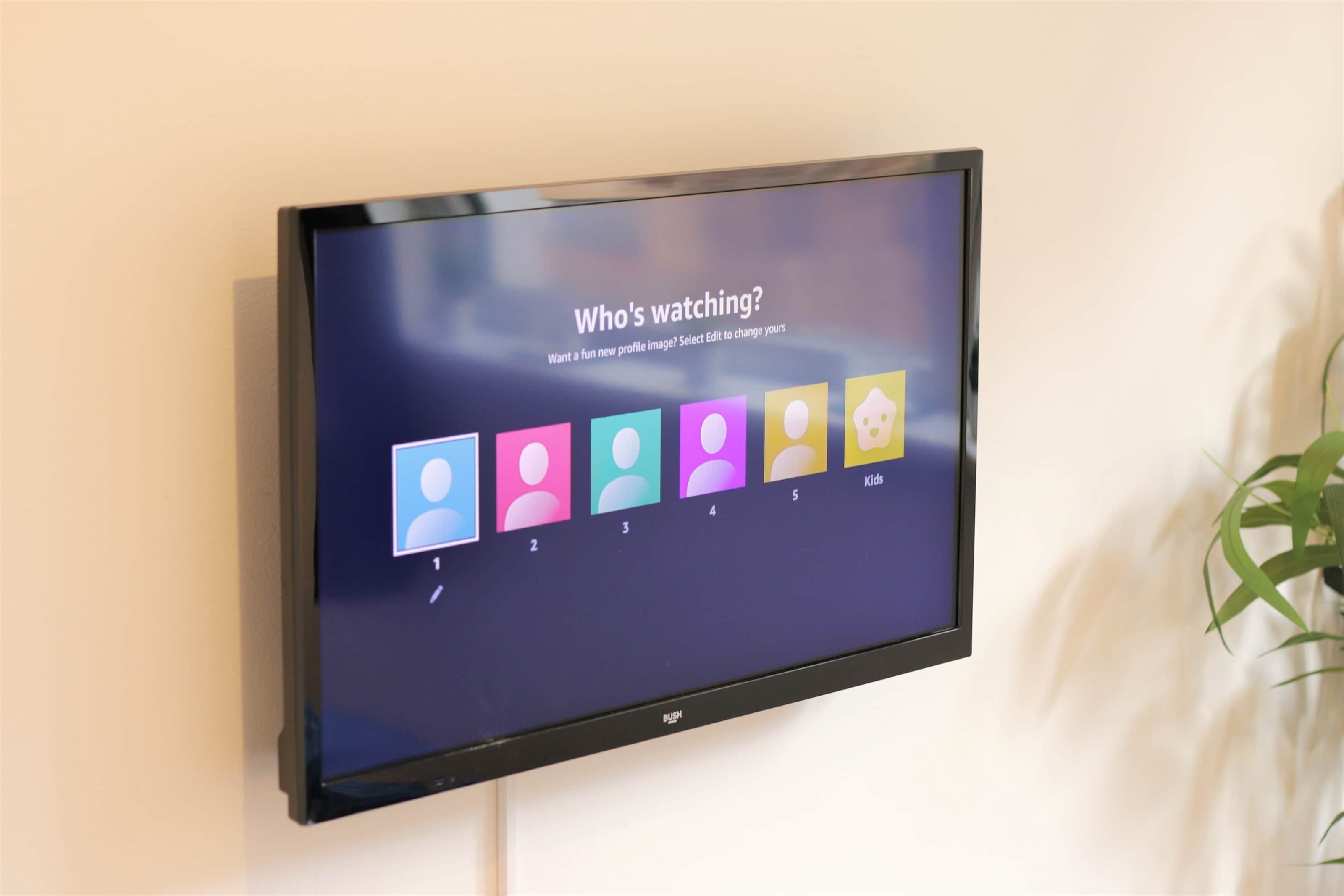 A flat-screen TV mounted on a wall displaying a social media interface with profile pictures and a 'Who's watching?' prompt.