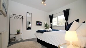 A bedroom with a bed, a bedside table with a lamp, a wall-mounted TV, a window with curtains, a hanging clothes rack, and a small plant on a shelf. - Ty Glas Mews (2 beds) (Cardiff)