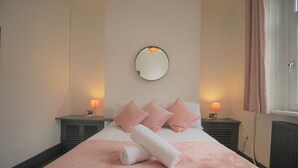 A neatly made bed with pink bedding and a rolled white towel, two bedside lamps, a round mirror, and a window with curtains. - Fitzroy Street (3 beds) (Cardiff)
