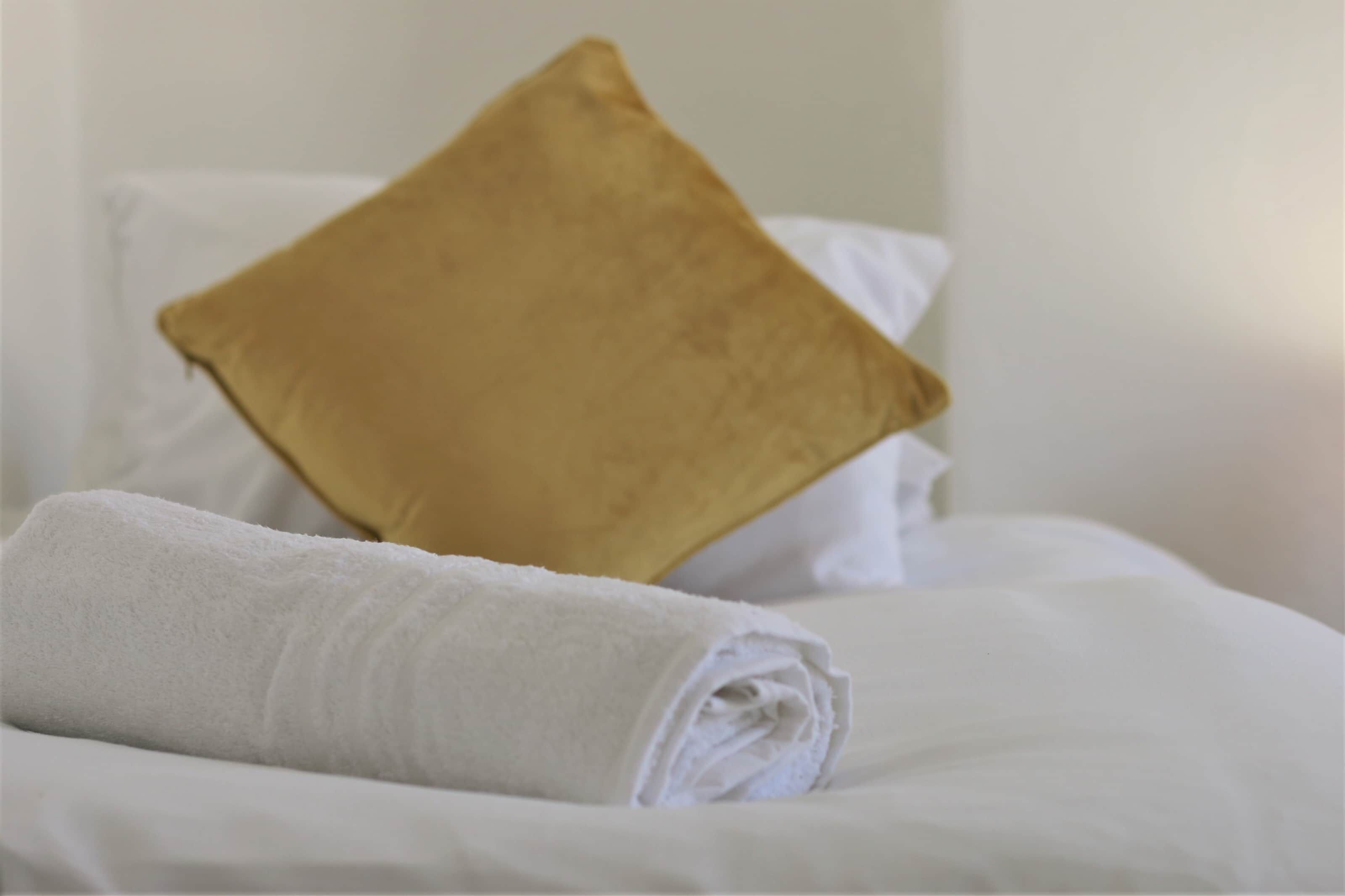 Una cama bien tendida con una toalla blanca enrollada y una almohada dorada.