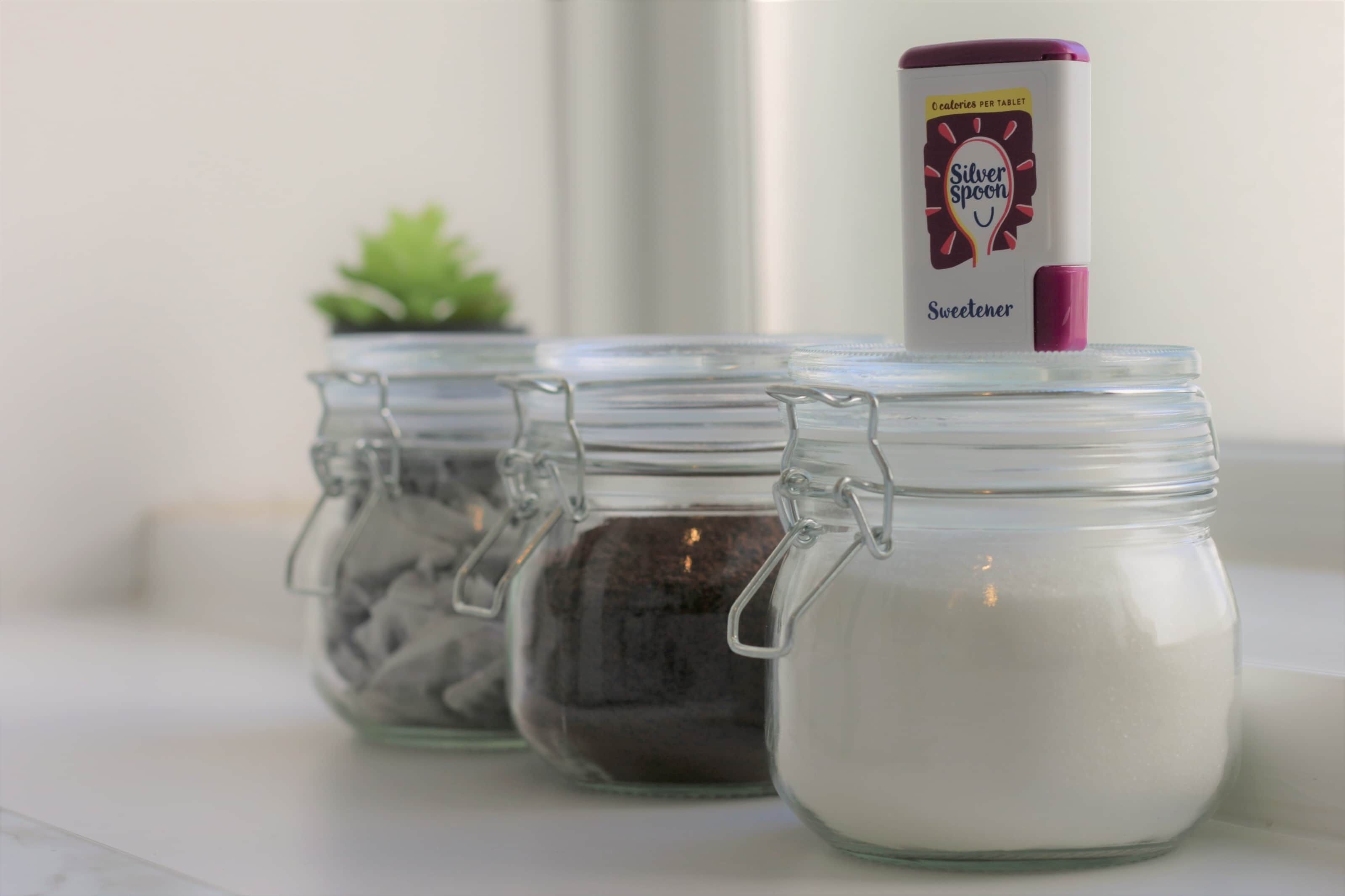 Three glass jars with metal clasps, one containing a white powder, another with a dark substance, and the third with a brownish material. A canister labeled "Silver Spoon Sweetener" is placed on top of the middle jar.