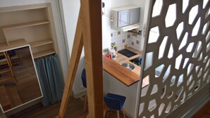 Eine kleine Küche mit Holzarbeitsplatte, Spüle und einem darüberliegenden Schrank. Im angrenzenden Raum steht ein blauer Stuhl und eine Holzleiter.
