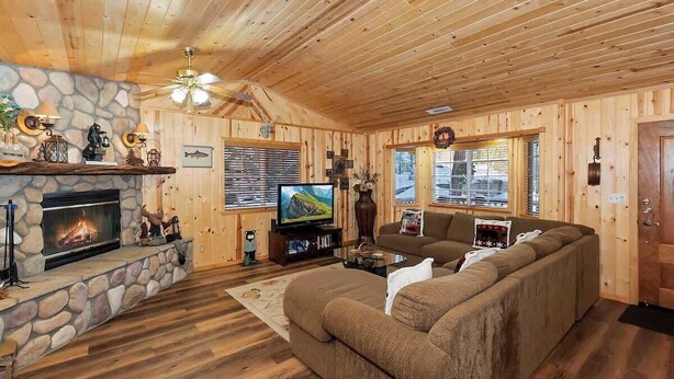 A cozy cabin interior with a stone fireplace, wooden walls, a large sofa, and a television. - Hibernation Station I Near Snow Summit (Big Bear Lake)