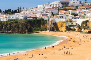 Una playa con gente tomando el sol y nadando, edificios blancos con techos anaranjados al fondo, y un acantilado rocoso.