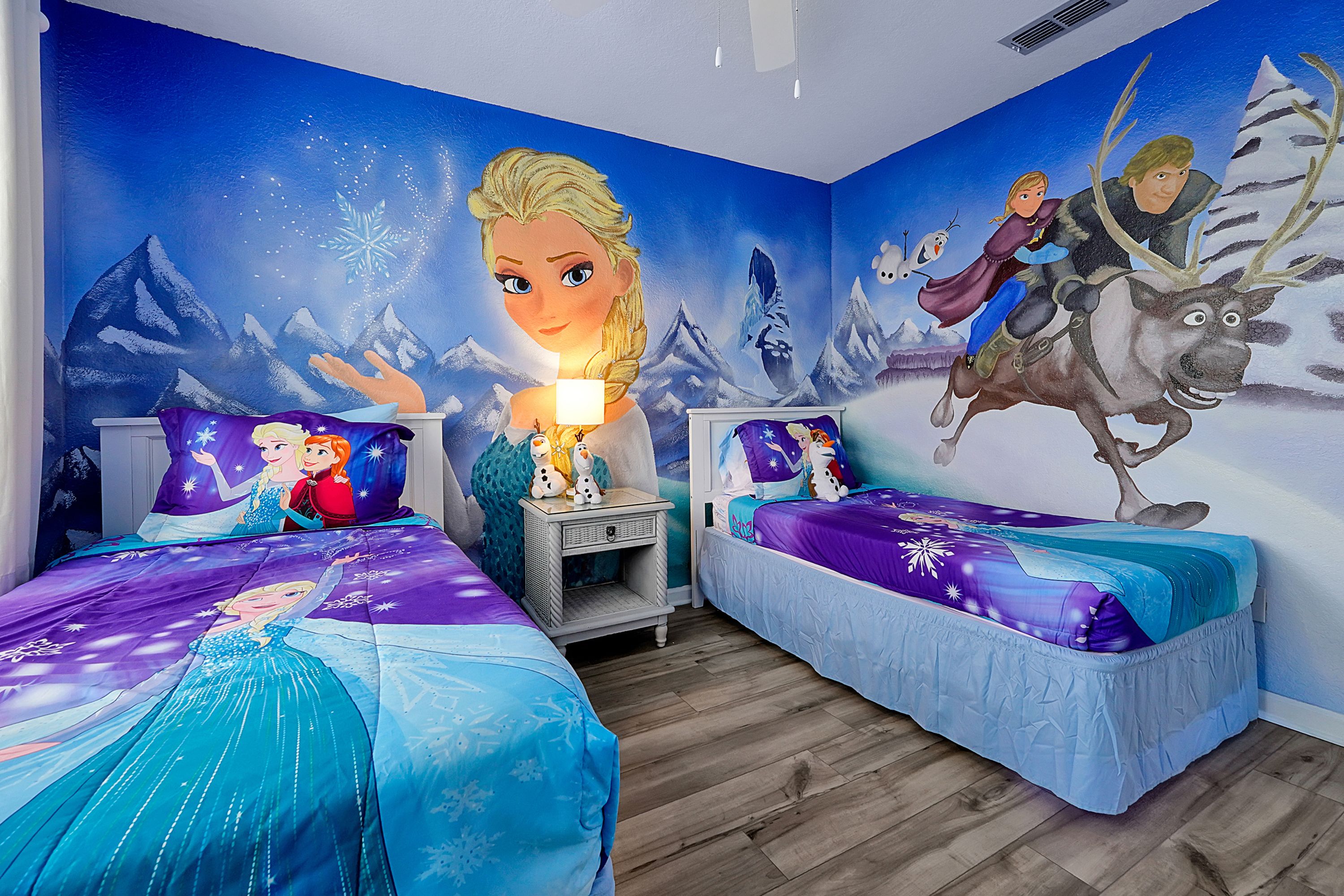 A bedroom with two beds, each with a themed bedspread, a nightstand with a lamp, and a mural on the wall depicting characters from a winter-themed movie.