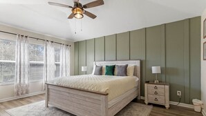 Un dormitorio con cama de madera, mesita de noche, lámpara, ventilador de techo y ventana con cortinas.
