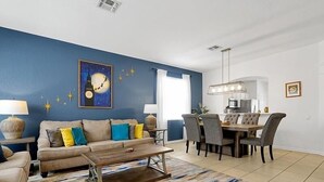 Una sala de estar con pared azul, una zona de comedor con mesa y sillas de madera, y una mesa baja con un tapete colorido.