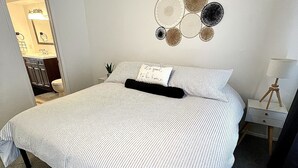 A neatly made bed with striped bedding and a pillow with text, a wall with circular wall art, a bedside table with a lamp, and a small potted plant. - Inner City TownHome (Fort Worth)