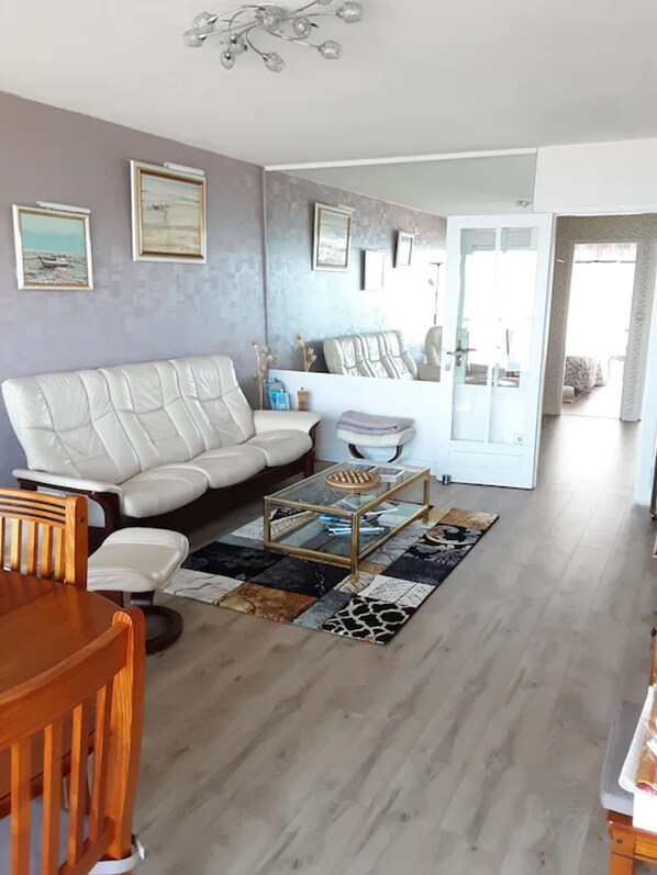 A living room with a white sofa, a glass coffee table, and a wooden dining table.