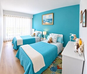 Une chambre avec deux lits, un bureau blanc et un tableau représentant une scène de plage.
