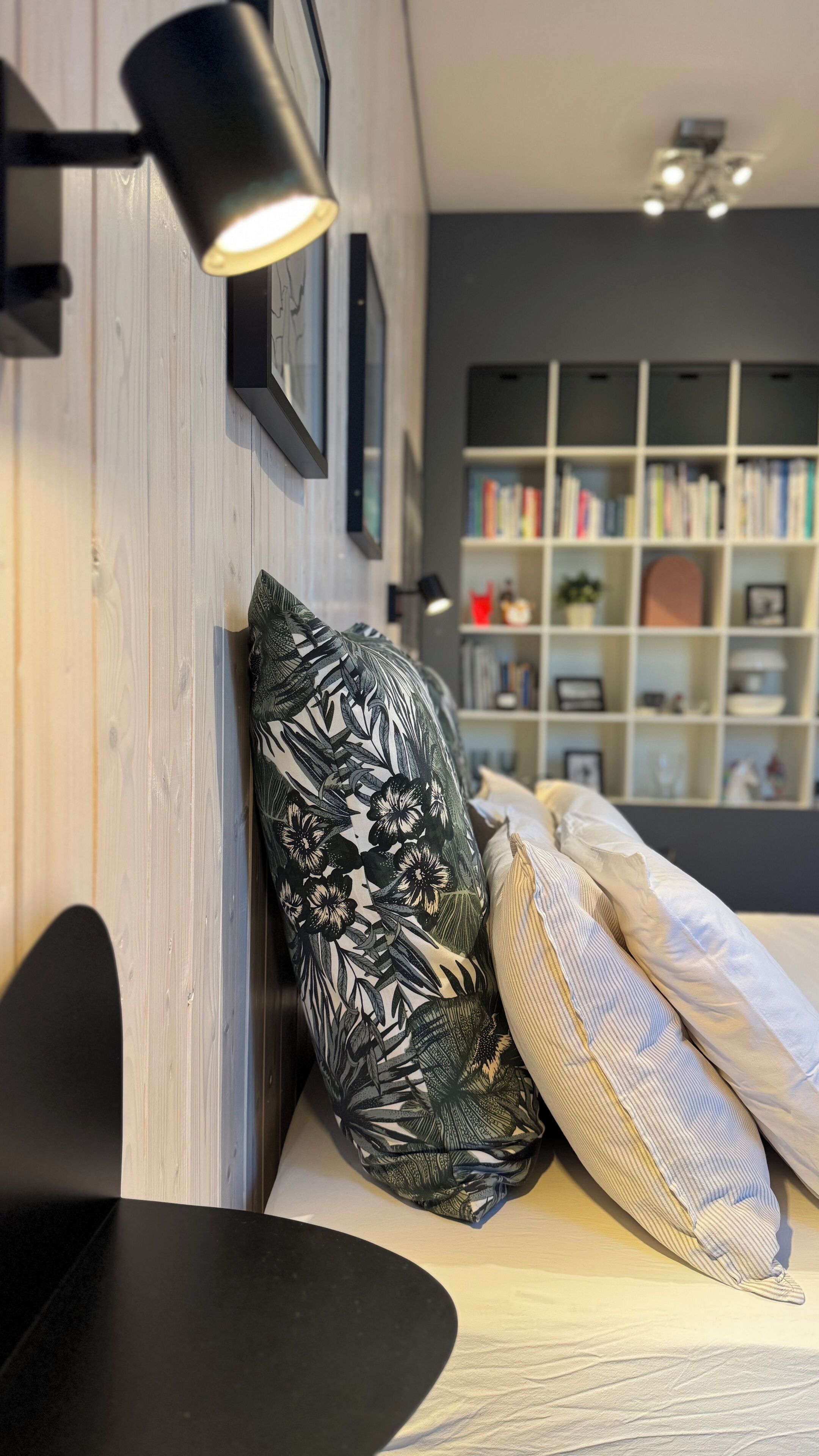 A modern room with a wooden wall, a black armchair, a patterned pillow, a striped pillow, and a bookshelf in the background.