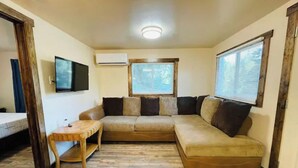 A cozy living room with a beige sectional sofa, a wooden coffee table, and a small side table. There is a window with blinds, an air conditioning unit, and a flat-screen TV mounted on the wall. - Mountain Bungalow (Martin City)