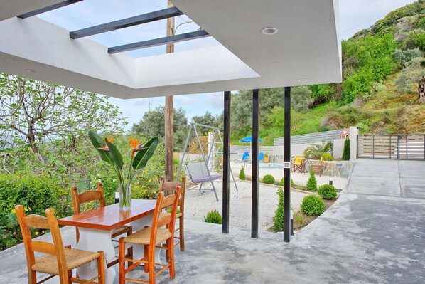 A covered patio area with a table and chairs, a swing, and a view of a pool and garden.