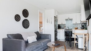 A modern living room with a grey sofa, a coffee table, and a kitchen area with white cabinets and a stainless steel refrigerator.