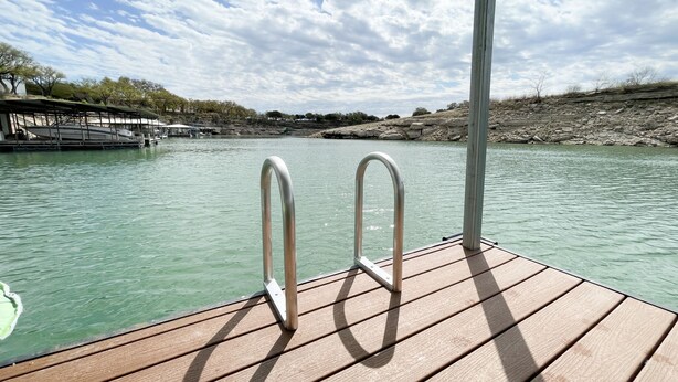 A wooden deck with metal railings overlooking a calm body of water, with a rocky shoreline and sparse vegetation in the background.