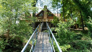 Eine Holzbrücke führt zu einer Hütte mit einem Steinkamin, umgeben von dichtem Grün.