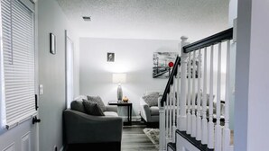 A hallway with a staircase leading to a second floor, a grey sofa, a small table with a lamp, and a framed picture on the wall.
