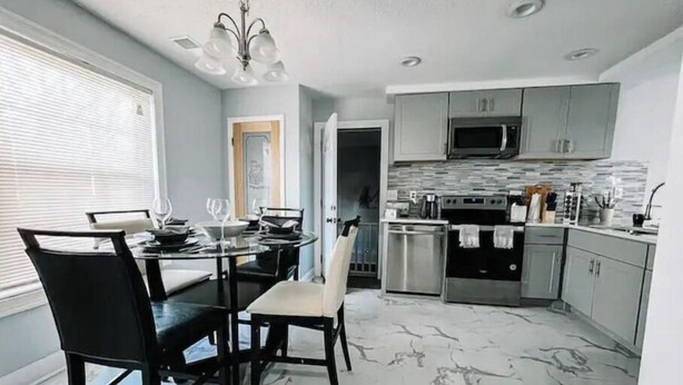 A modern kitchen with stainless steel appliances, a dining area with a glass table and chairs, and a light-colored marble floor.