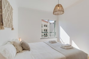 A bedroom with a bed, a macrame wall hanging, a hanging light fixture, and a window with a view of buildings.